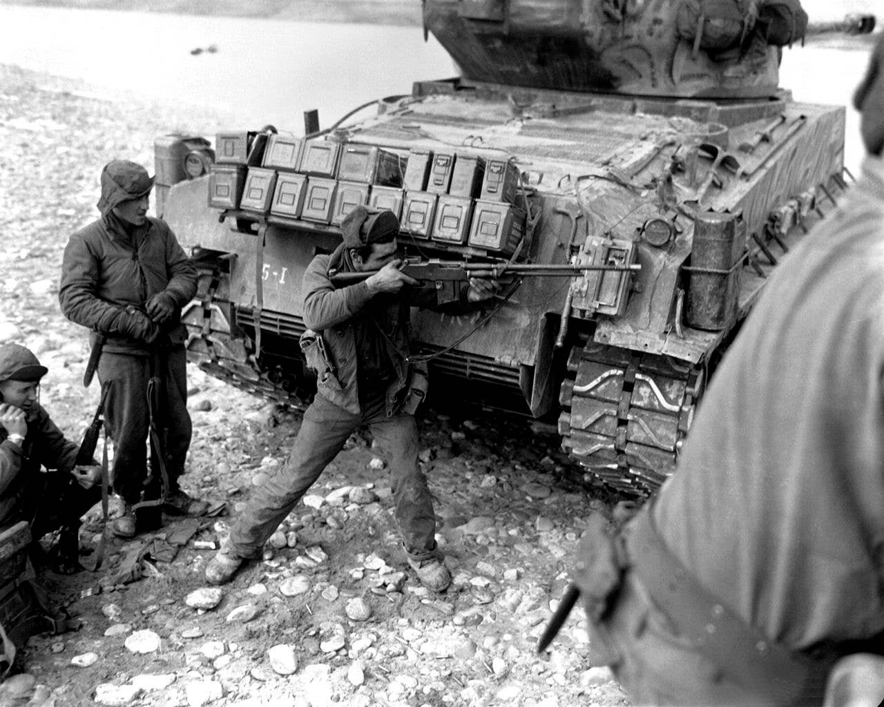 Taking cover behind their tank escort, one man of this ranger patrol of the 5th RCT, U.S. 24th Infantry Division, uses his BAR to return the heavy Chinese Communist small arms and mortar fire which has them pinned down on the bank of the Han River. At left another soldier uses a field radio to report the situation to headquarters. (Wikimedia Commons)