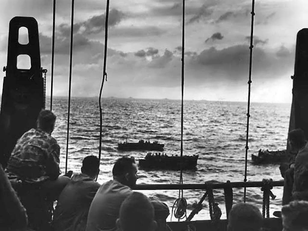 Little suspecting of the hell that awaits them, U.S. Marines wait aboard a Coast Guard manned combat transport at Tarawa for the invasion barges that will take them ashore. Beyond the rail Coast Guard coxswains can be seen maneuvering loaded barges. On the horizon other vessels are faintly visible. (Public domain)