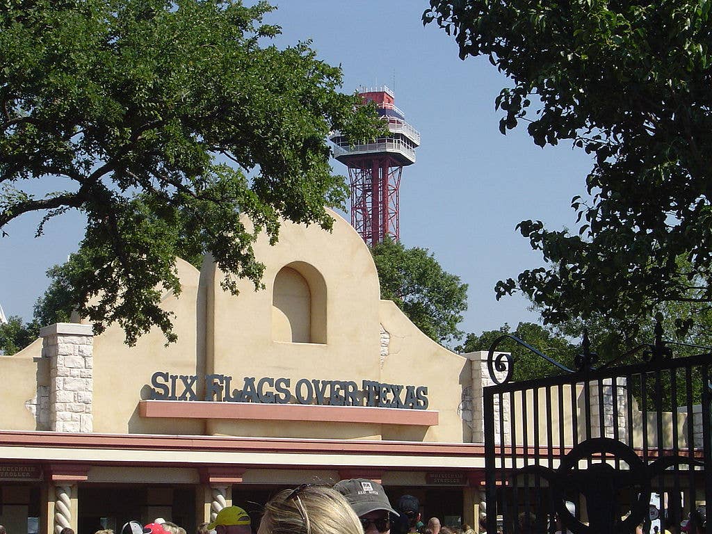 The entrance of Six Flags over Texas welcomes visitors while the Oil Derrick <a href="https://en.wikipedia.org/wiki/Observation_tower">observation tower</a> looms in the background.
