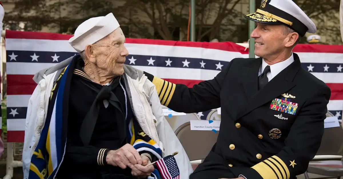 An older retired sailor stands next to a younger sailor in uniform.
