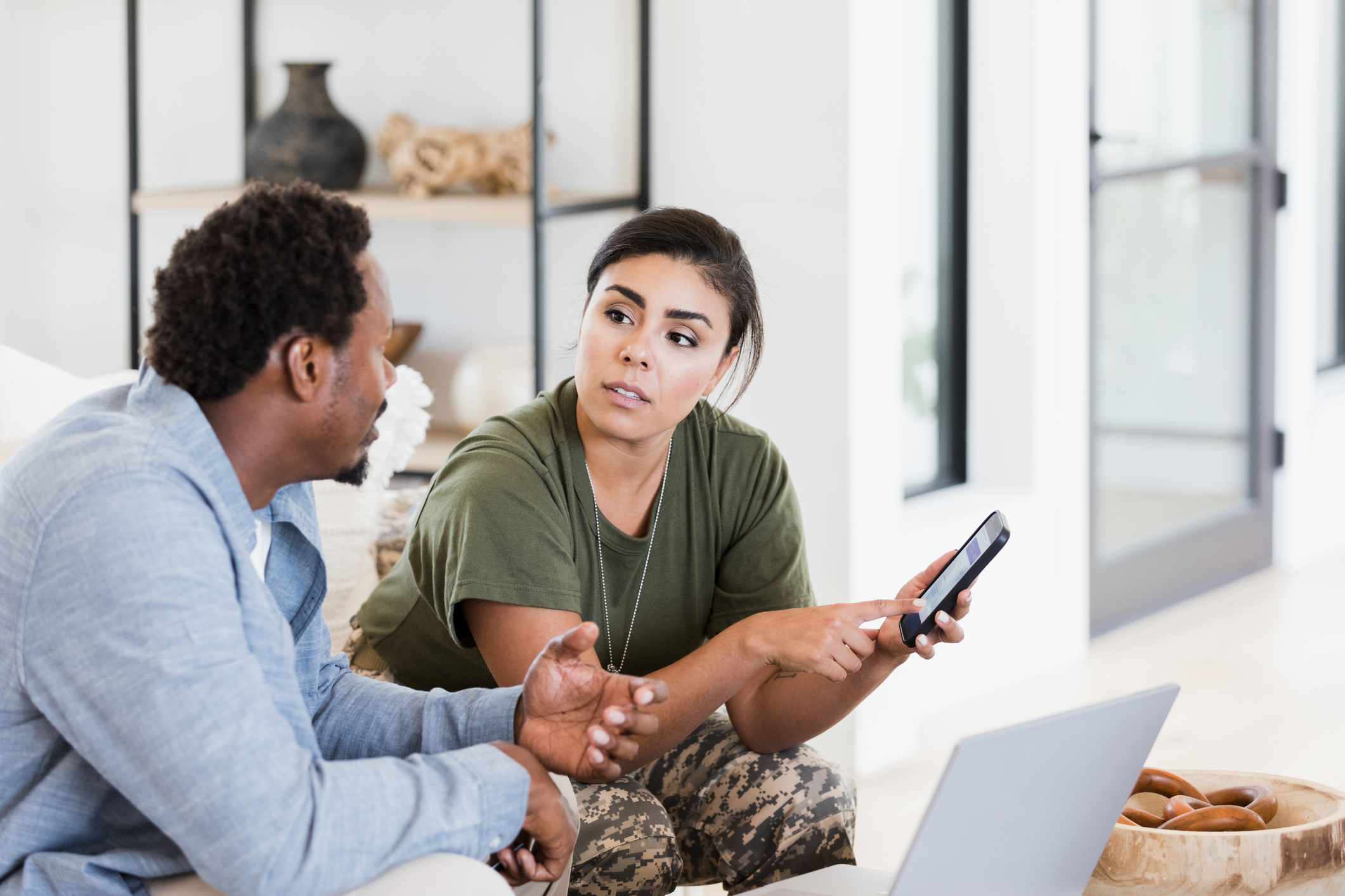 Sitting in their living room, the mid adult man gestures as he talks to his mid adult soldier wife about their home finances or an online purchase.  She is pointing to something on the smart phone screen and they have their laptop on the coffee table in front of them.