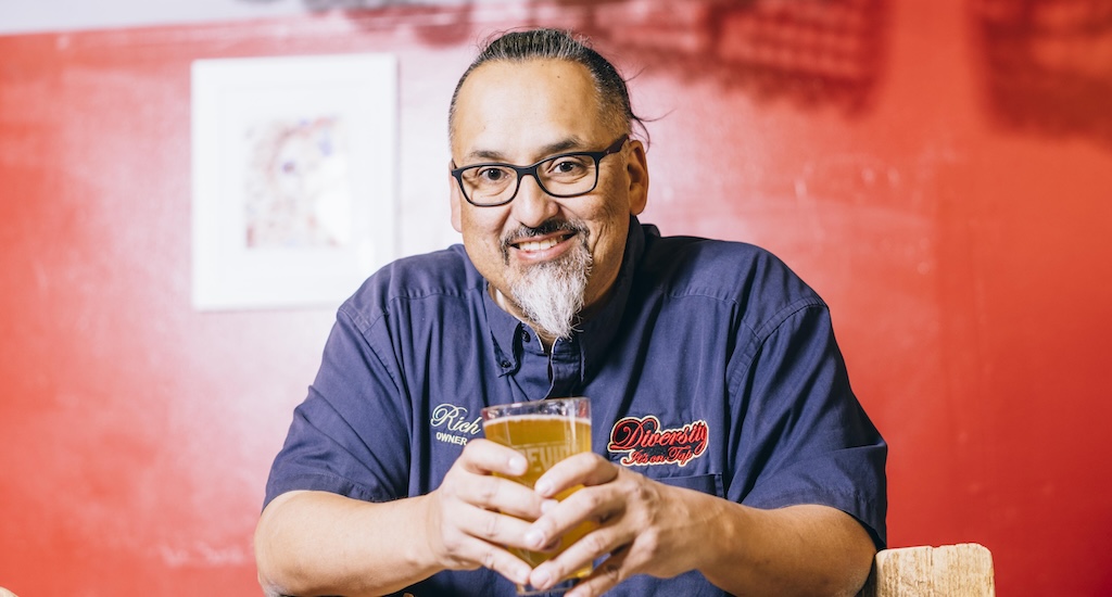 Richard Fierro standing in front of a red wall and holding a beer.
