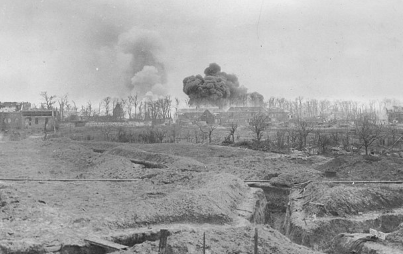A series of trenches in a French war zone during World War I; smoke is billowing from several buildings which have been damaged by bombs or fire.