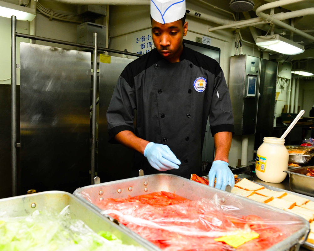 a Navy soldier makes sandwiches with bread, mayonnaise, lettuce and tomatoes. 