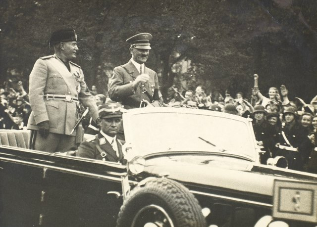 Adolph Hitler and Benito Mussolini ride in a convertible with crowds watching during WWII