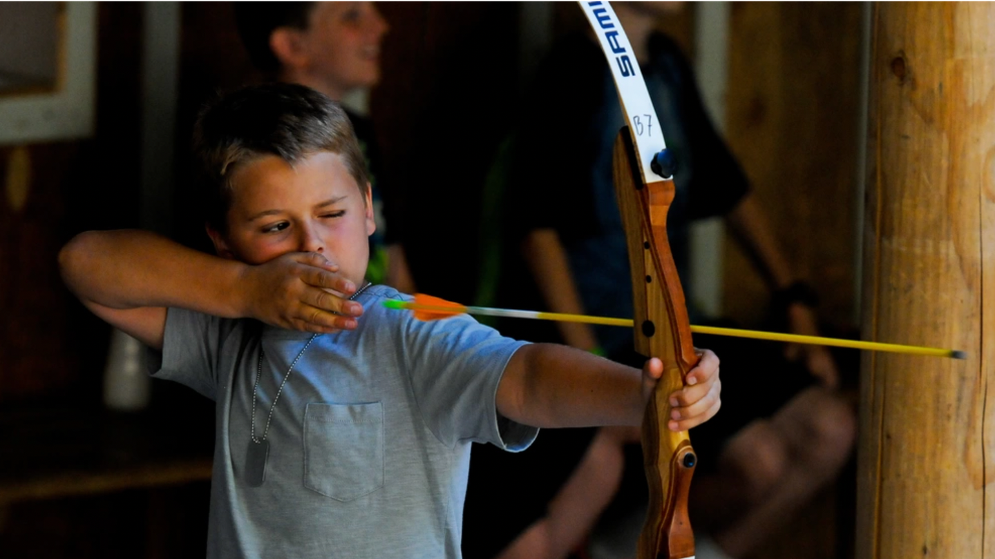A boy aims a bow and arrow at summer camp