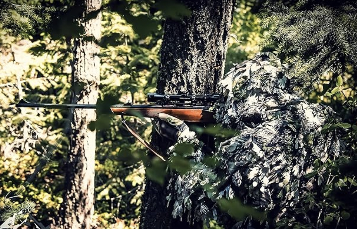 A hunter points their gun in the woods while wearing a ghillie suit