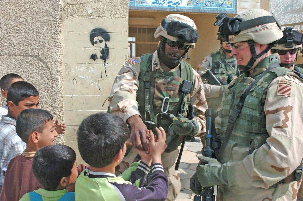 Soldiers from B Co., 3/15 Infantry hand out hard candy to kids in Sadr City, Iraq, Feb. 28, 2003. An ominous stencil of Shia cleric Muqtada al-Sadr looms in the background.