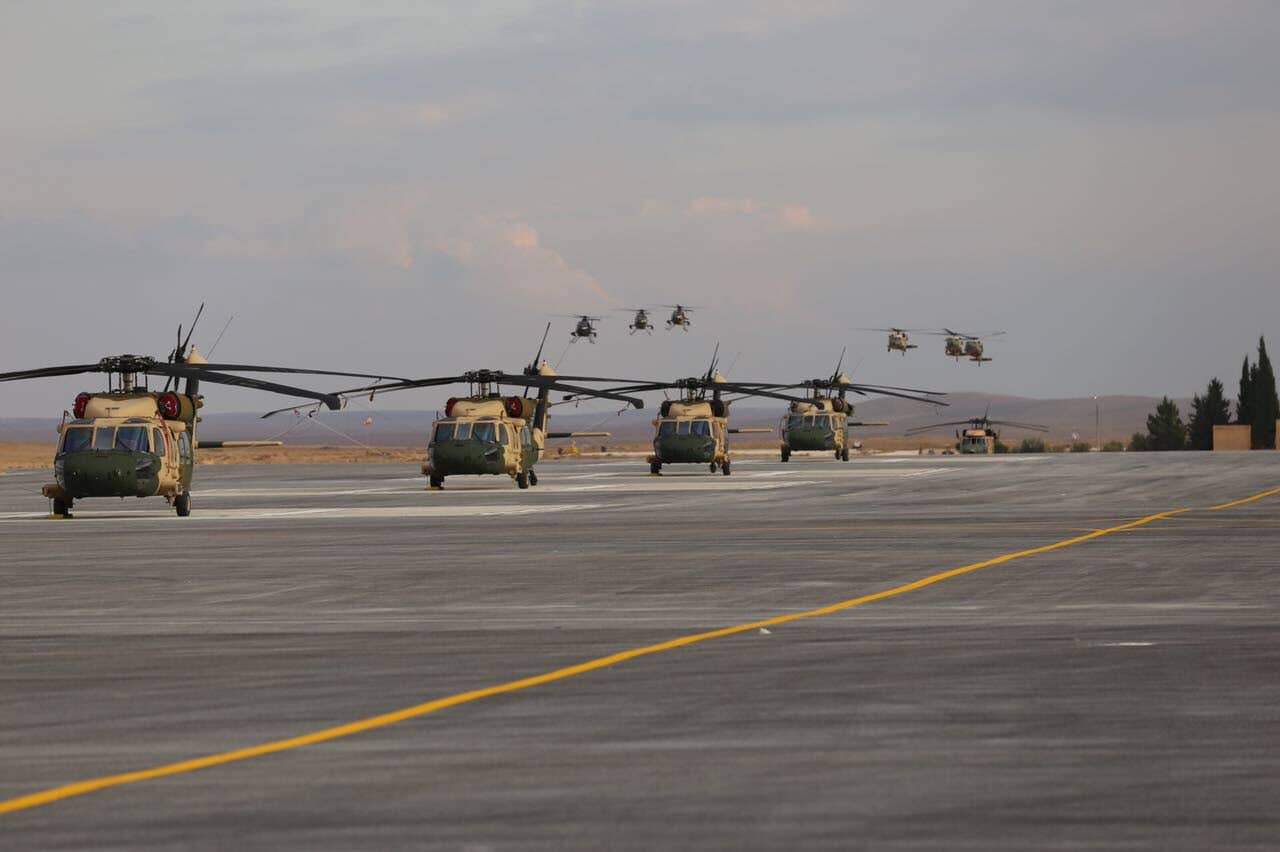 UH-60 Black Hawks are on the tarmac while a mix of UH-60s and MD530Fs fly overhead. (U.S. State Department photo)