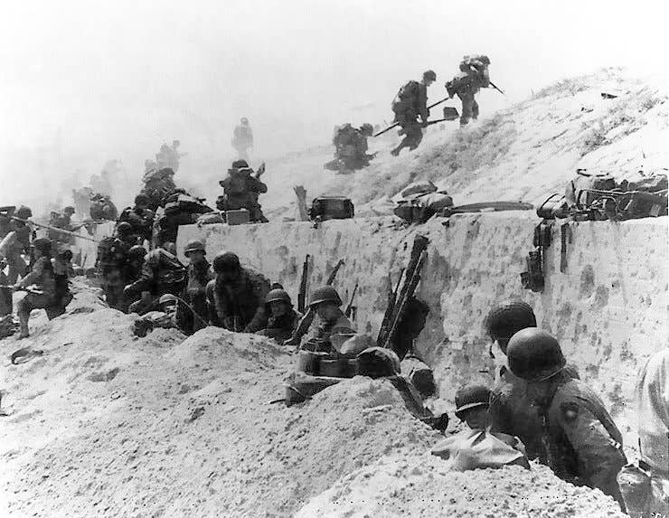 U.S. Soldiers of the 8th Infantry Regiment, 4th Infantry Division, move out over the seawall on Utah Beach after coming ashore. Other troops are resting behind the concrete wall. (Army Signal Corps photo)