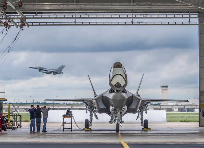 Big and Little Brother: An F-35A sits in a run station on the Fort Worth, Texas, flight line, while an F-16 Fighting Falcon, also produced at the Fort Worth plant, takes off in the background. Learn more about F-35 production. | Lockheed Martin photo