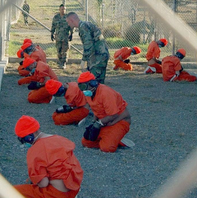 Detainees in orange jumpsuits sit in a holding area under the watchful eyes of Military Police at Camp X-Ray at Naval Base Guantanamo Bay, Cuba, during in-processing to the temporary detention facility on Jan. 11, 2002.(DoD photo by Petty Officer 1st class Shane T. McCoy, U.S. Navy)