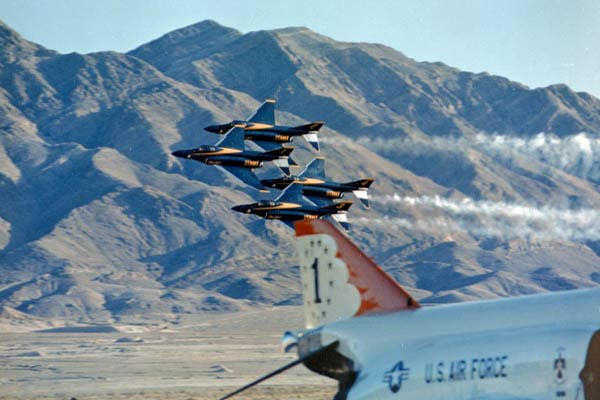 Blue Angels diamond flies along show center at Nellis AFB with Thunderbird No. 1 parked on the ramp in the foreground. (Photo: U.S. Navy)