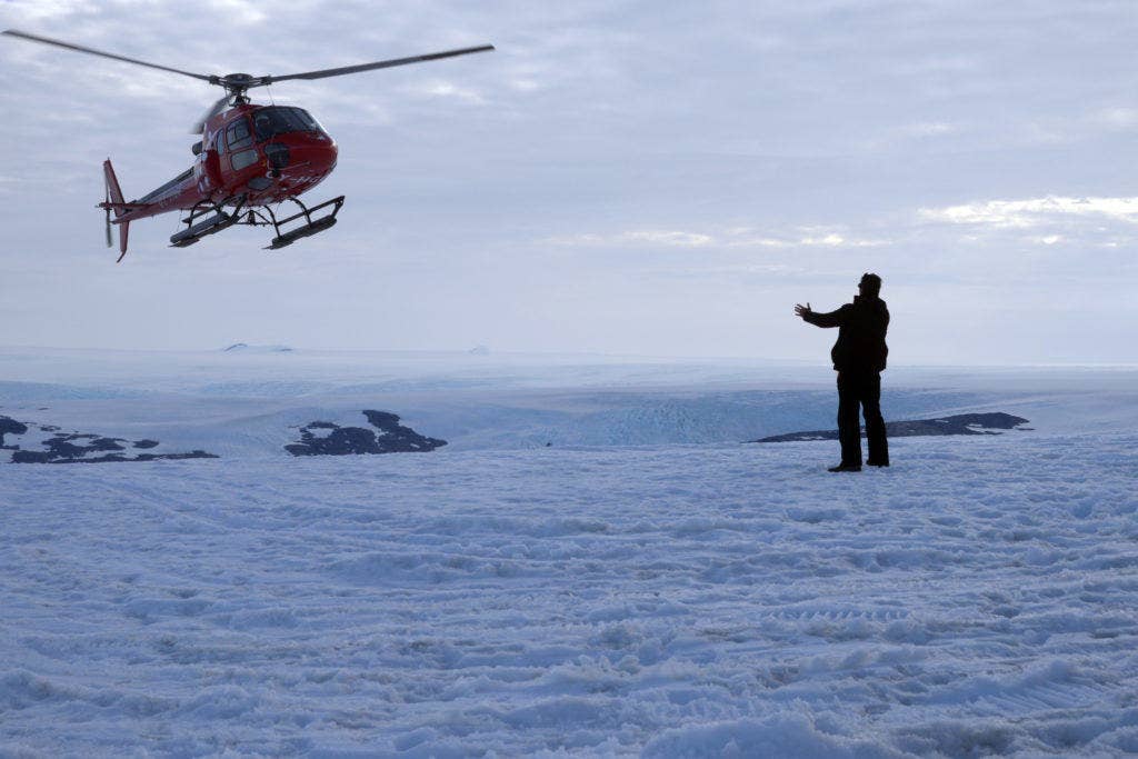 Mitchell Zuckoff, an author embedded with the Joint Recovery Mission - Greenland, signals to helicopter pilot Tom Andreassen, of Air Greenland, where to land near the nunatak on a glacier near Koge Bay, Greenland, Aug. 16, 2013. (U.S. Coast Guard photo by Petty Officer 2nd Class Jetta H. Disco.)