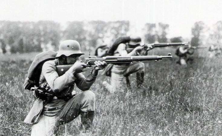 Soldiers crouch in a field, using a precursor weapon to the M16