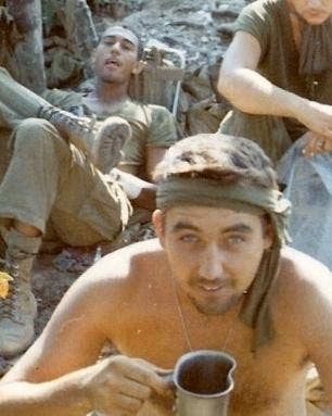 Bob Gunton, shirtless, holds a drink while other soldiers look on