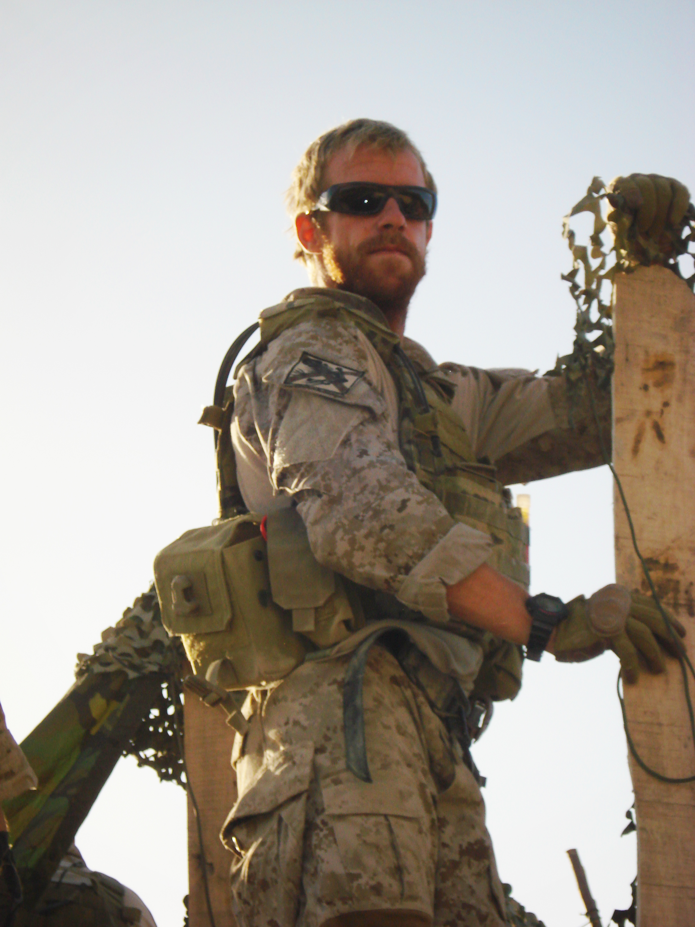 A soldier wearing desert camouflage uniform, tactical vest, gloves, and sunglasses stands holding a wooden post wrapped with camouflage netting. The soldier has a beard and a watch on the left wrist, with a tan pouch attached to the vest. The background is a clear sky with bright sunlight.