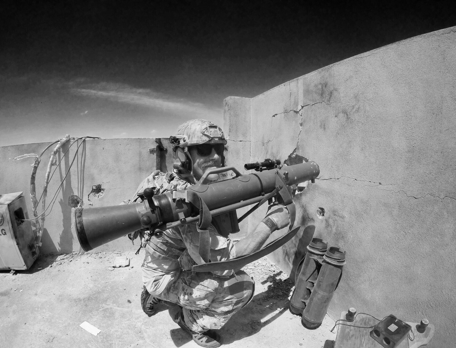 A soldier in camouflage uniform and helmet is crouching while aiming a rocket launcher. The scene is outdoors with a cracked concrete wall behind him, showing bullet holes. Nearby on the ground are three rocket launcher tubes and some electronic equipment with wires. The image is in black and white.