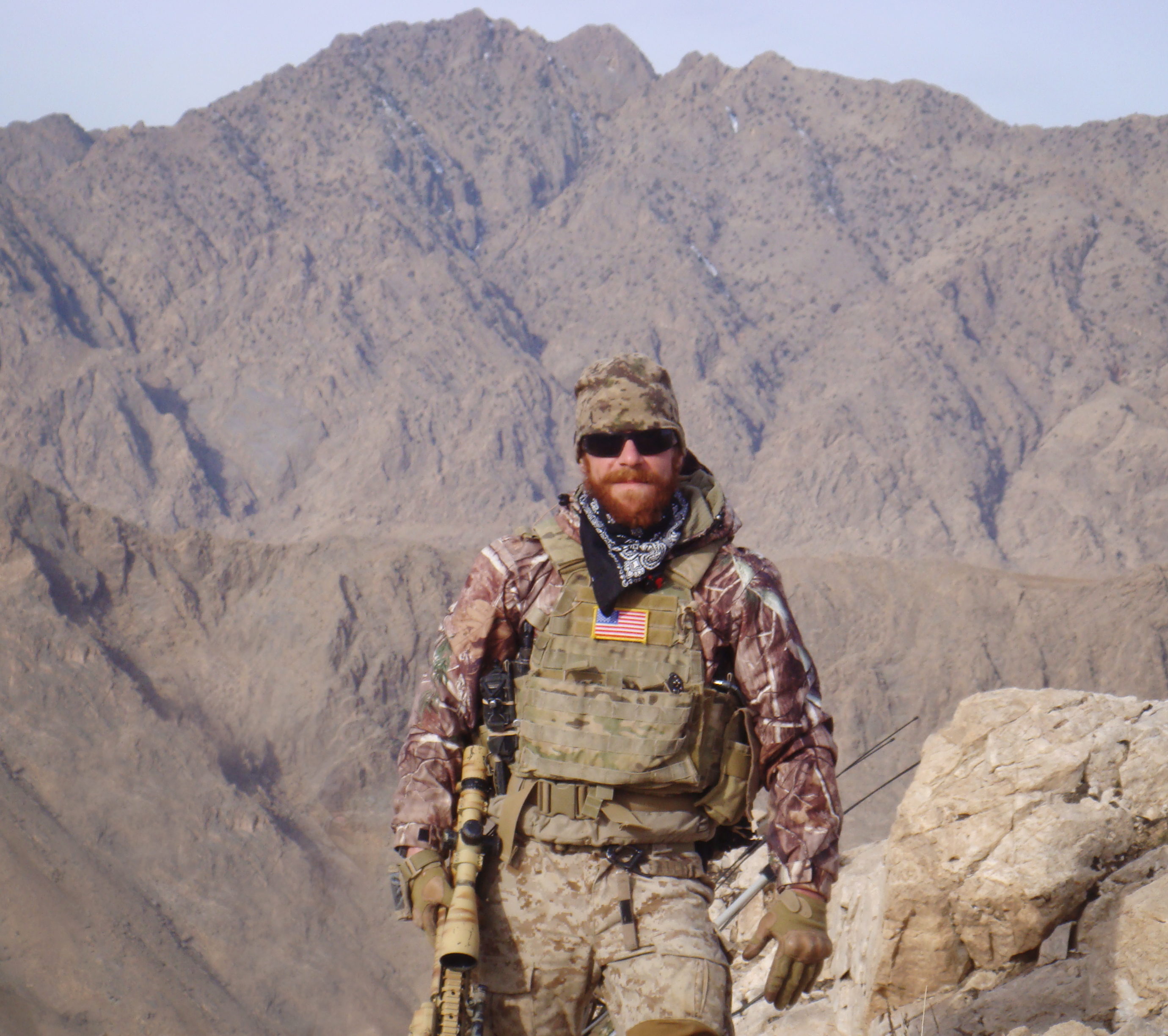 A soldier wearing desert camouflage uniform, tactical vest with an American flag patch, gloves, and sunglasses stands in front of a rocky mountainous background. The soldier has a beard, a bandana around the neck, and is holding a rifle.