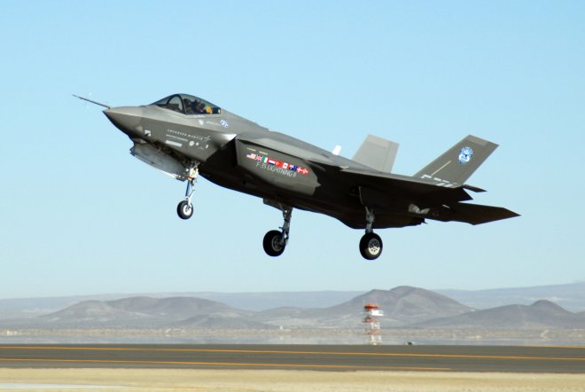 A gray F-35 Lightning II fighter jet is captured mid-air just above the runway during takeoff or landing. The aircraft has its landing gear extended and displays multiple national flags and the text "F-35 LIGHTNING II" on the side. The background shows a clear blue sky and distant mountains.