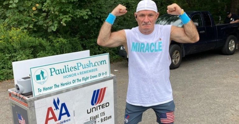 Paulie Veneto wearing a white sleeveless shirt with the word "MIRACLE" in blue, blue wristbands, a white backward cap, and blue shorts with an American flag patch is flexing his arm muscles. He stands next to a sign that reads "PauliesPush.com HONOR & REMEMBER To Honor The Heroics Of The Flight Crews Of 9-11" with logos for American Airlines and United Airlines and flight details for Flight 11, Flight 77, Flight 175, and Flight 93. There is a black pickup truck and greenery in the background.