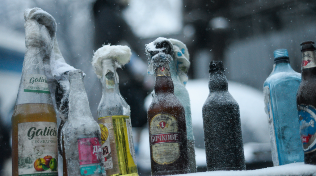 Several glass bottles covered in frost and snow are lined up outdoors in a cold, snowy environment. The bottles vary in color and label design, with some having caps wrapped in snow or ice. The background is blurred, emphasizing the frozen bottles in the foreground.