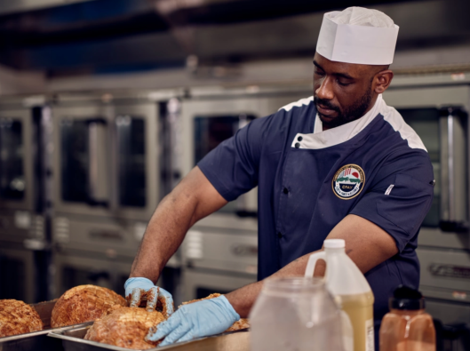 A chef wearing a white hat, navy blue uniform with a patch, and blue gloves is handling freshly baked bread loaves on a tray in a commercial kitchen. Various containers and bottles are visible in the foreground, with industrial ovens in the background.