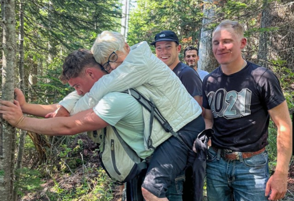 A group of five people in a forest setting. One person, an older woman with short white hair and glasses, is being carried on the back of a younger man wearing a light-colored shirt and backpack, who is holding onto a tree for support. Behind them, three men stand close together, smiling. One man is wearing a black cap and dark shirt, another has a light blue shirt, and the third is wearing a black t-shirt with "2021" printed on it and jeans. The scene is shaded by trees with sunlight filtering through.