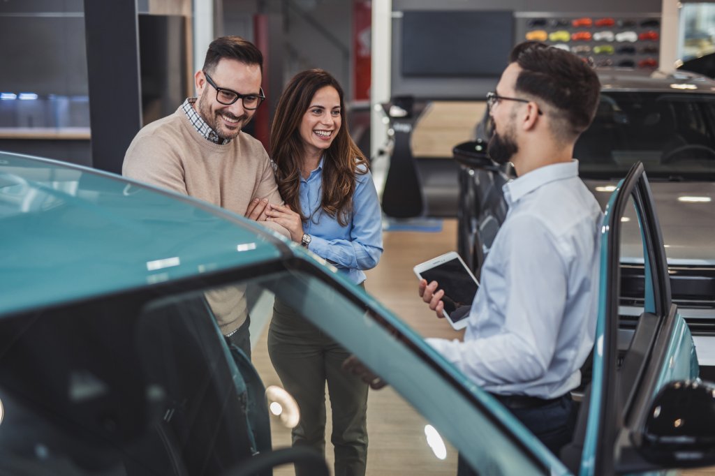 Happy couple holding hands and smiling while listening to the car salesman presenting details about a new car in a vibrant dealership showroom, enjoying the purchasing experience together