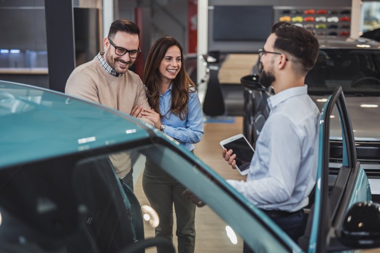 Happy couple holding hands and smiling while listening to the car salesman presenting details about a new car in a vibrant dealership showroom, enjoying the purchasing experience together