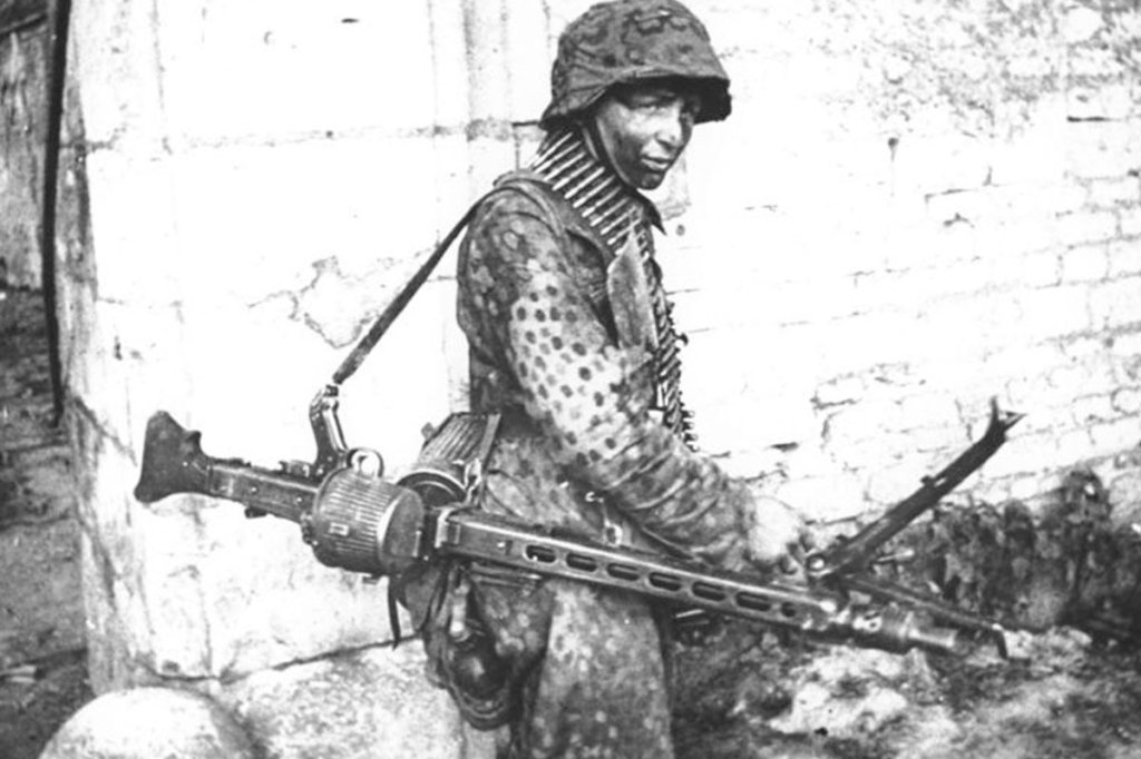 A Waffen-SS soldier in 1944 carrying an MG 42 configured as a light support weapon with a folding bipod and 50-round basket drum containing the ammo belt.