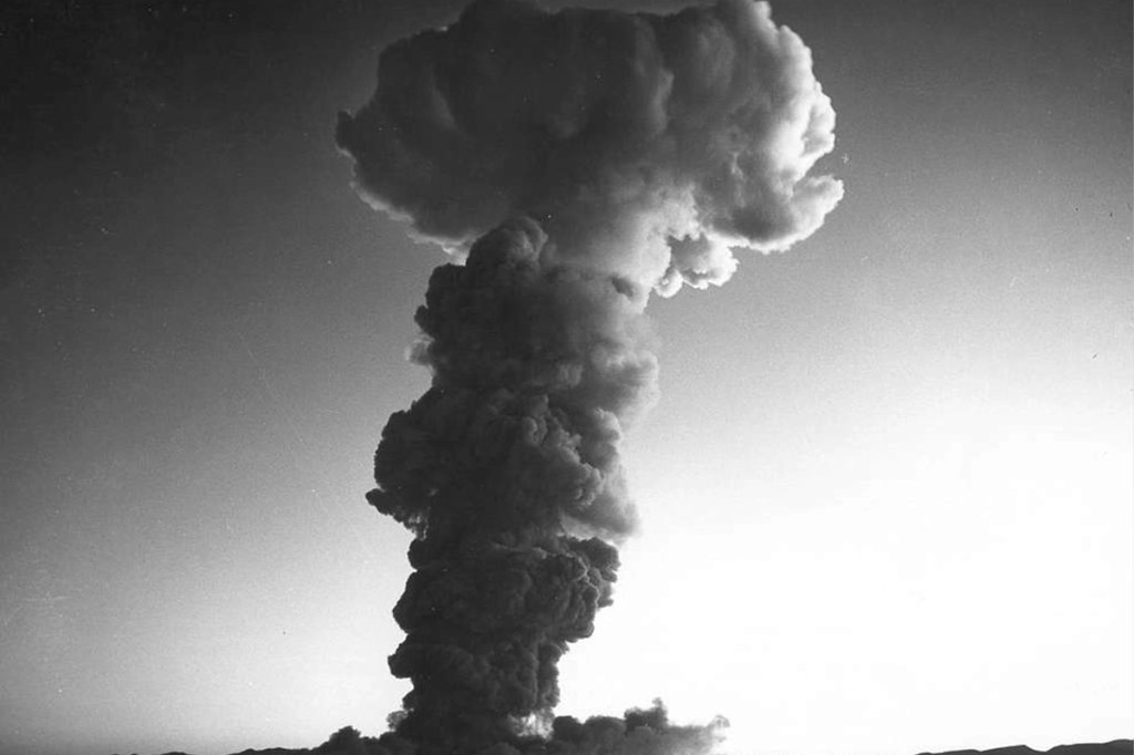 mushroom cloud begins to form above Yucca Flat. The July 5, 1957 above nominal shot sent a thermal wave across the desert, igniting bushes and other growth on nearby foothills near the surface of the ground.