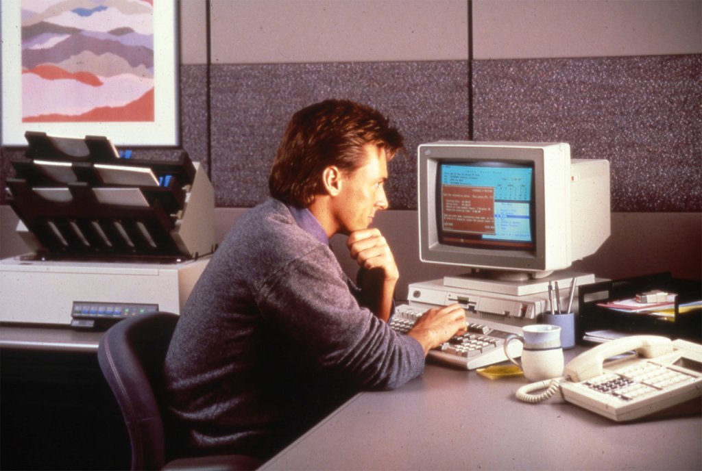 A man in a casual shirt and nice haircut stares pensively into the monitor of a PROFS (Professional Office System) PS2 Model 30