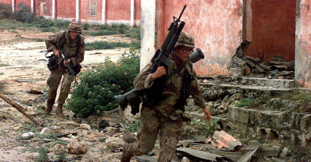 Two US Marines run toward the camera around a dilapidated building. The Marine in the foreground carries a M16A2 rifle with the M203 Grenade Launcher attached. Slung over his shoulder he has a M136 (AT4) light anti-armor weapon. The Marine in the background carries a M16A2 rifle pointed toward the ground. They are running for cover as sniper shots are fired from undisclosed areas of the cantonment area. The Marines are from Task Force Mogadishu. Their mission is in direct support of Operation Restore Hope.