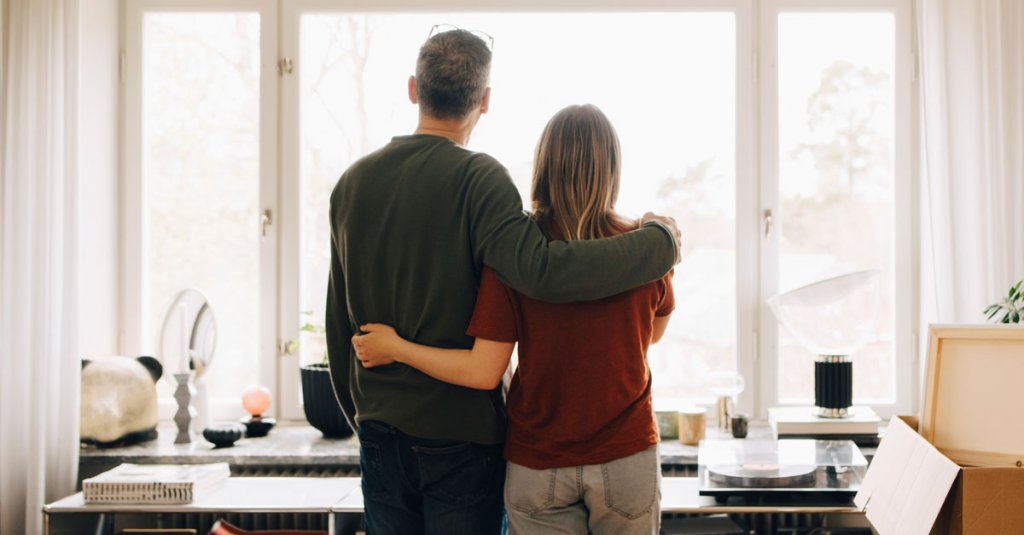 Rear view of father and daughter standing with arms around while looking through window at home