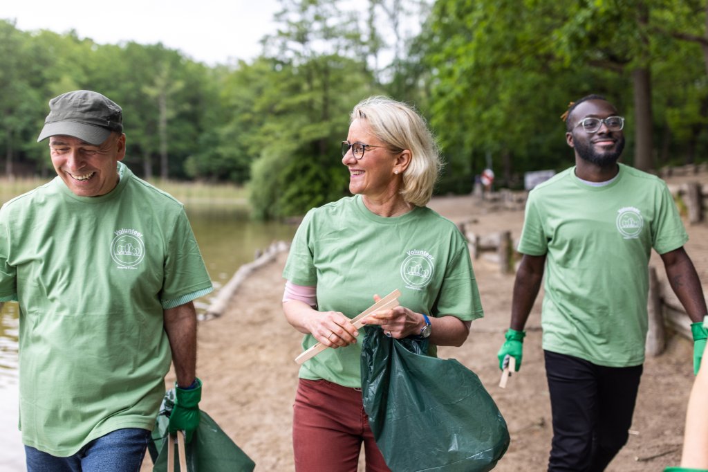 Mature man and woman with volunteers cleaning lakeside. Group of community service people in green t-shirts walking along the beach with garbage bags to collect the waste.