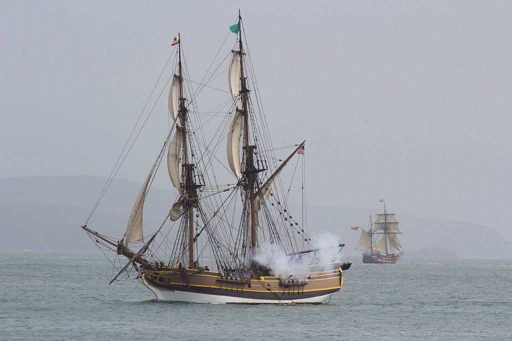 Two sailing ships hold a mock battle with the shoreline distantly visible behind them.