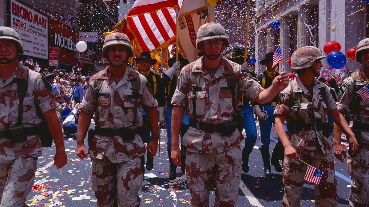 Soldiers marching in the Desert Storm Victory parade with flags and confetti behind them.