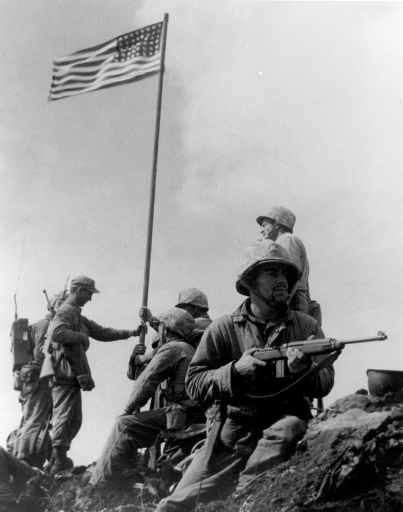 A Marine holding a M1 carbine covers personnel during the first flag-raising on Mount Suribachi.